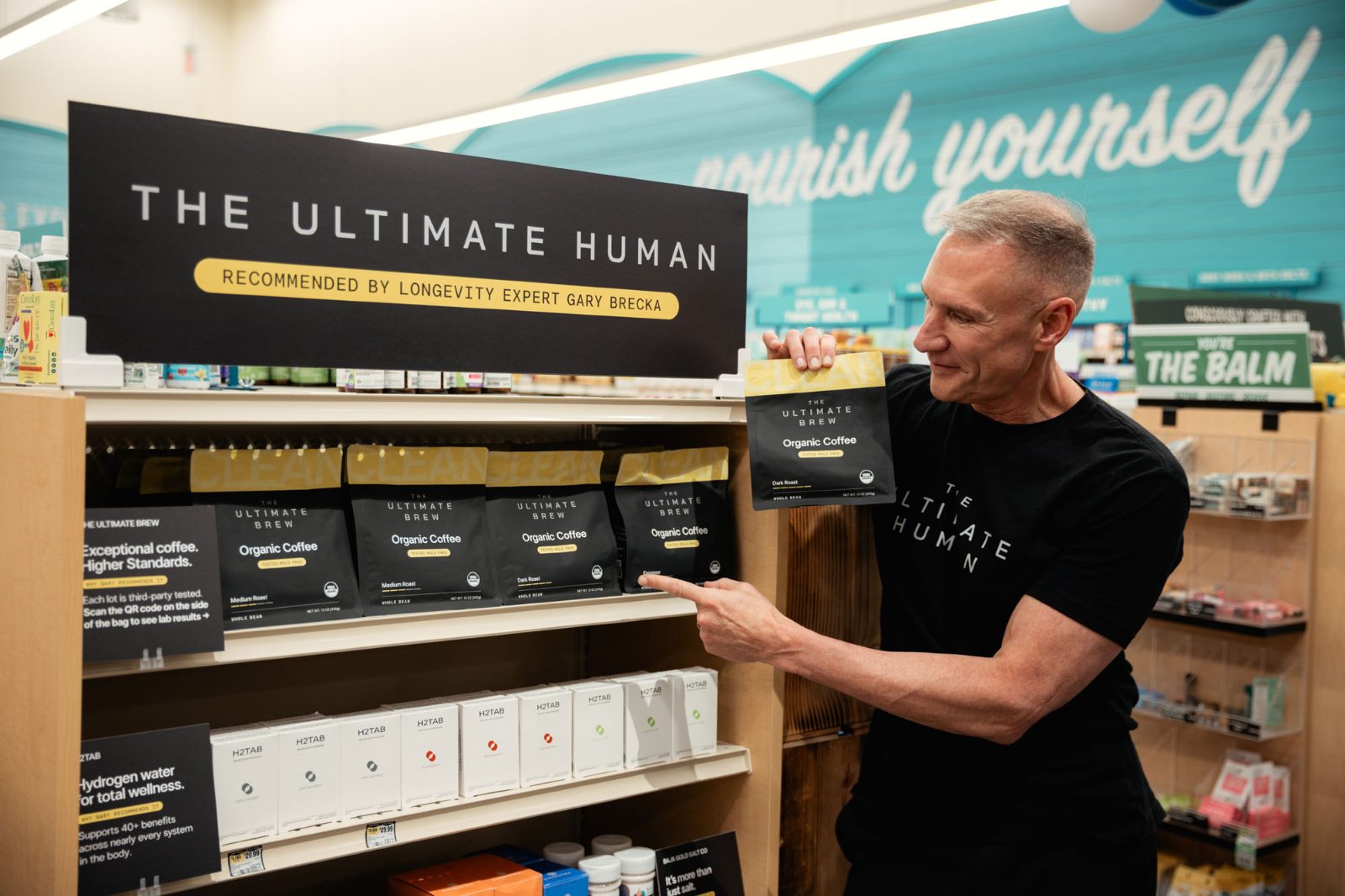 Gary Brecka holds a bag of The Ultimate Brew and points at the coffee shelf of a Sprouts endcap